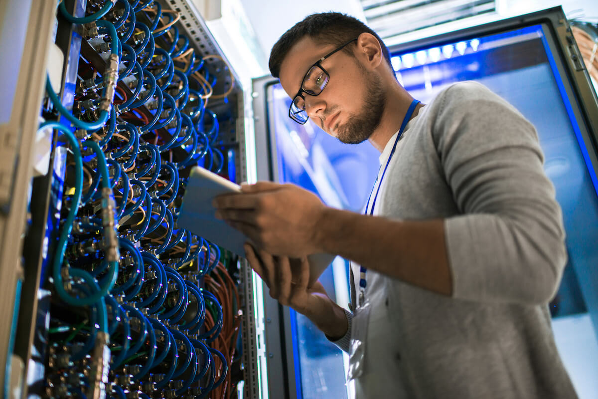 A network engineer inspecting cables in a server room while using a tablet.
